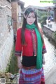 A woman in a red shirt and green scarf standing by a river.